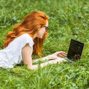 Une jeune femme rousse avec des lunettes noires, travaillant sur un ordinateur portable, allongée dans l'herbe