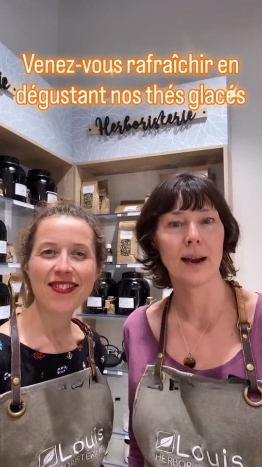 Juliette et Carole souriantes en tablier Louis Herboristerie à Saint-Ouen-sur-Seine, devant des étagères de plantes médicinales avec l'inscription textuelle sur les thés glacés.