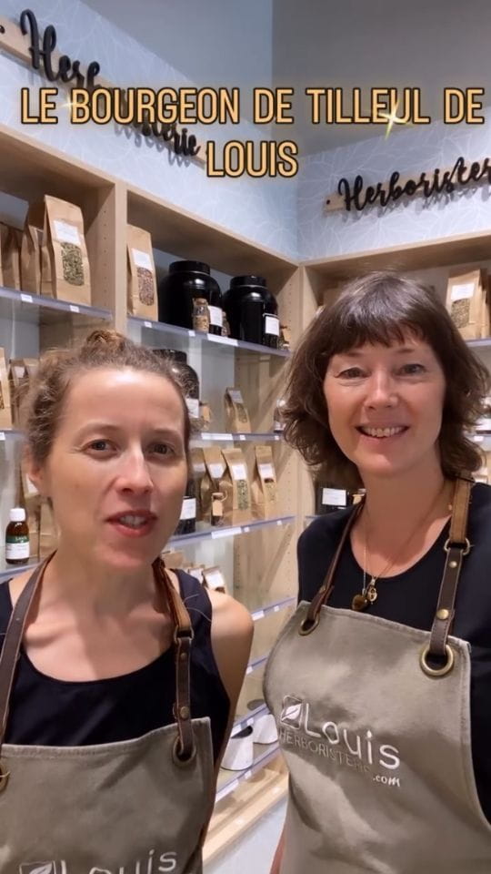 Deux femmes souriantes en tablier Louis Herboristerie devant des étagères de sachets de plantes, surmontées du texte LE BOURGEON DE TILLEUL DE LOUIS.
